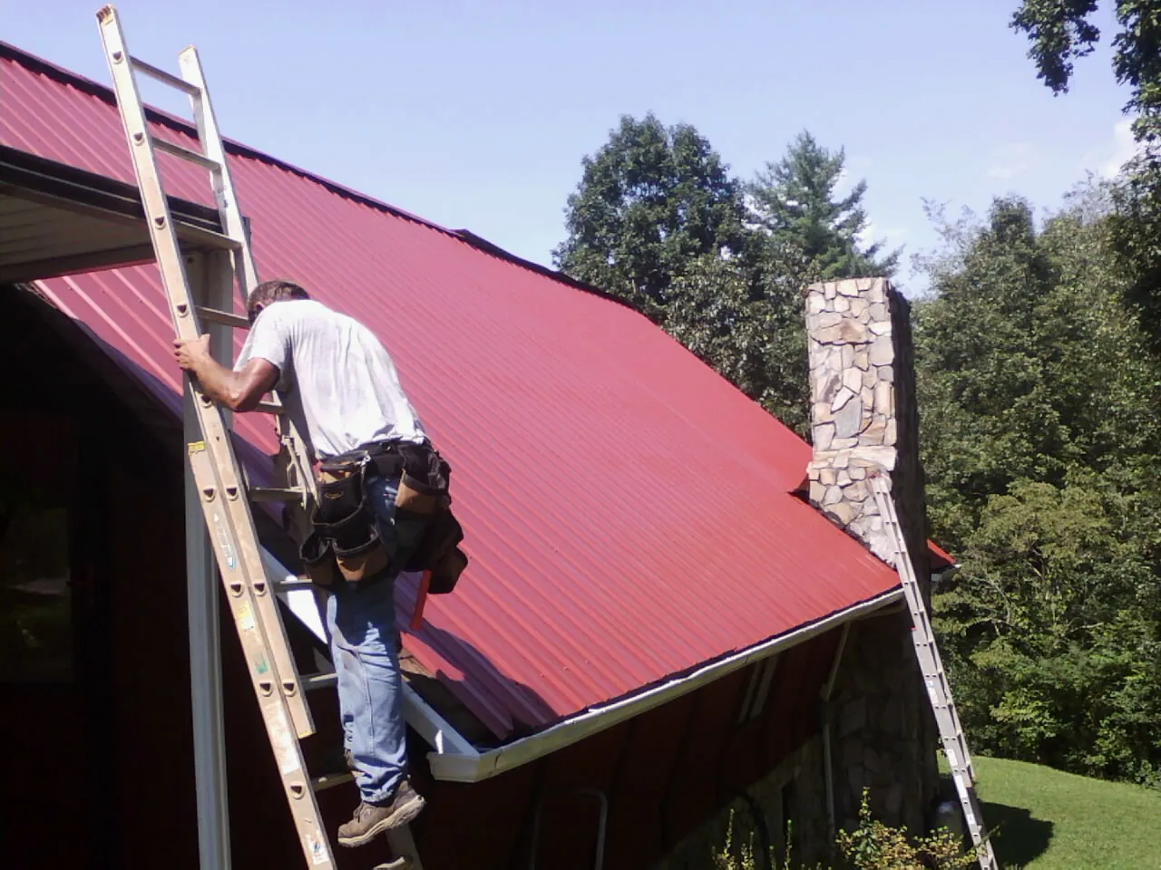 Roofer on a ladder beside a red metal roof and stone chimney — Soffit & Fascia Repair by Roofing Brothers in Camden Point